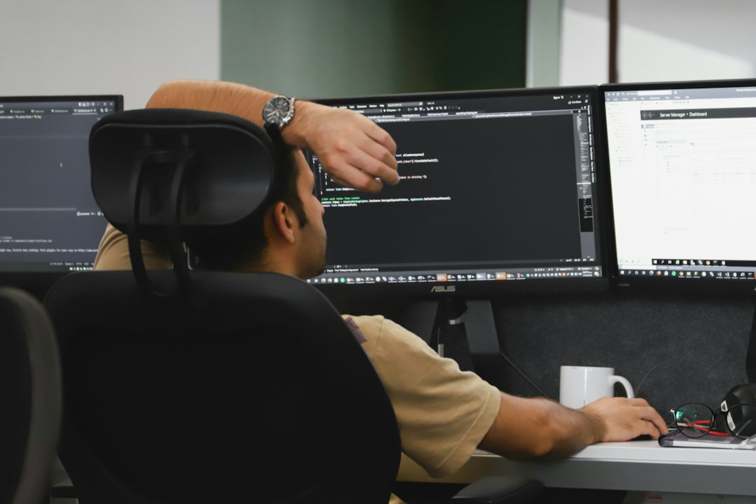 Man seated at a desk coding on three monitors displaying dark-theme code, with a white mug and glasses on the desk nearby.