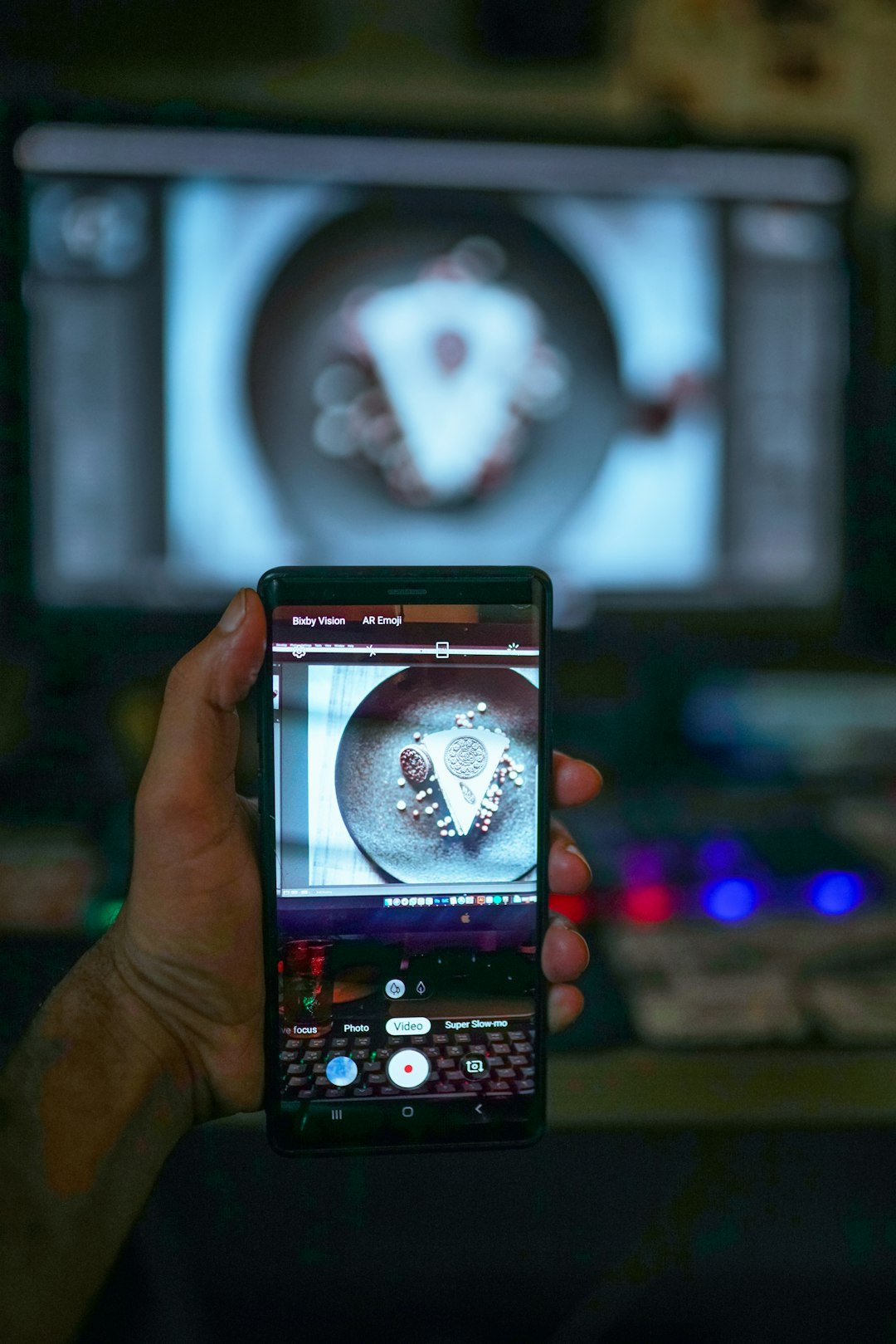 Person holds a phone and photographs a round plate with a triangular slice of pizza topped with pepperoni/sauce in a dim room.