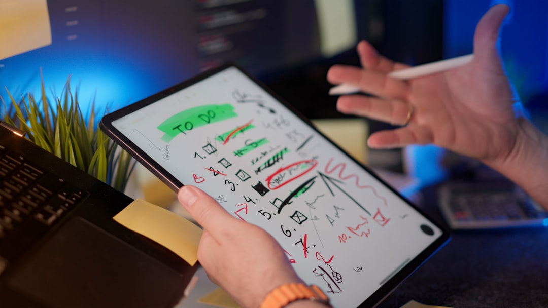 Tablet screen shows a handwritten 'TO DO' list with checkboxes and doodles, held by a person gesturing over a desk with a sticky note and plant nearby.