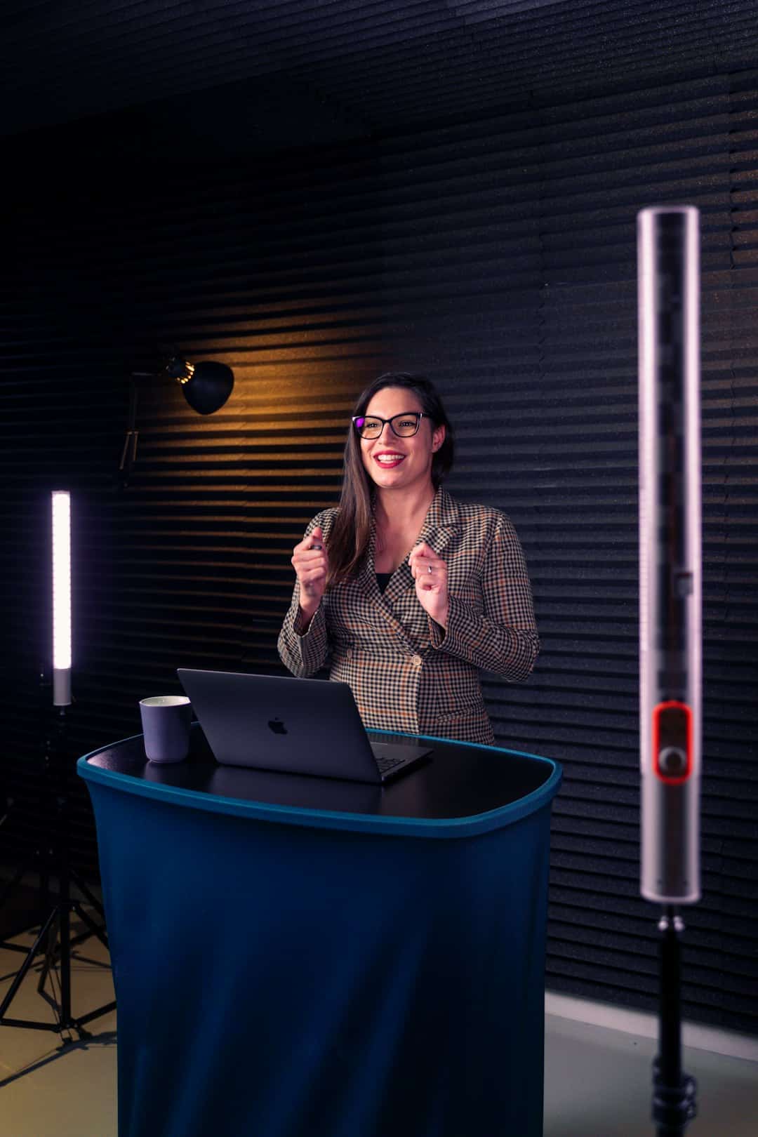 A woman in a checkered blazer and glasses stands behind a blue podium with an open laptop, presenting in a recording studio.