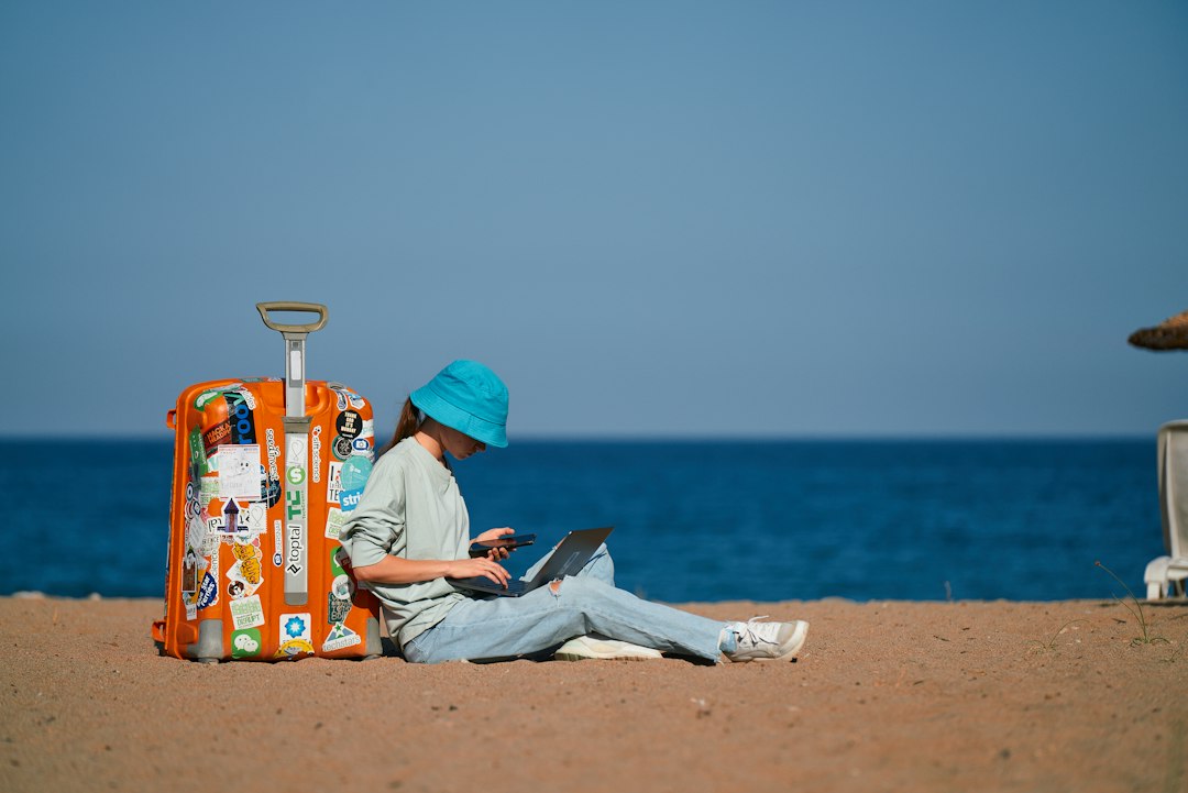 Traveler sits on the sandy beach beside an orange sticker-covered suitcase, using a laptop and a phone with the ocean in the background.