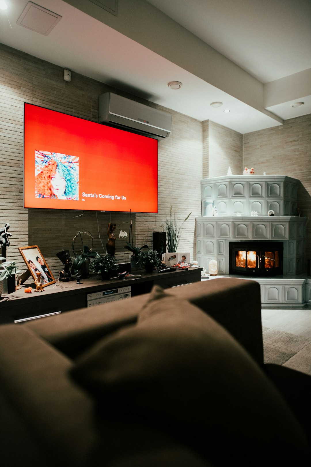 Living room with a large wall-mounted red TV above a low cabinet full of photos and plants, against a stone wall, in a cozy lounge.