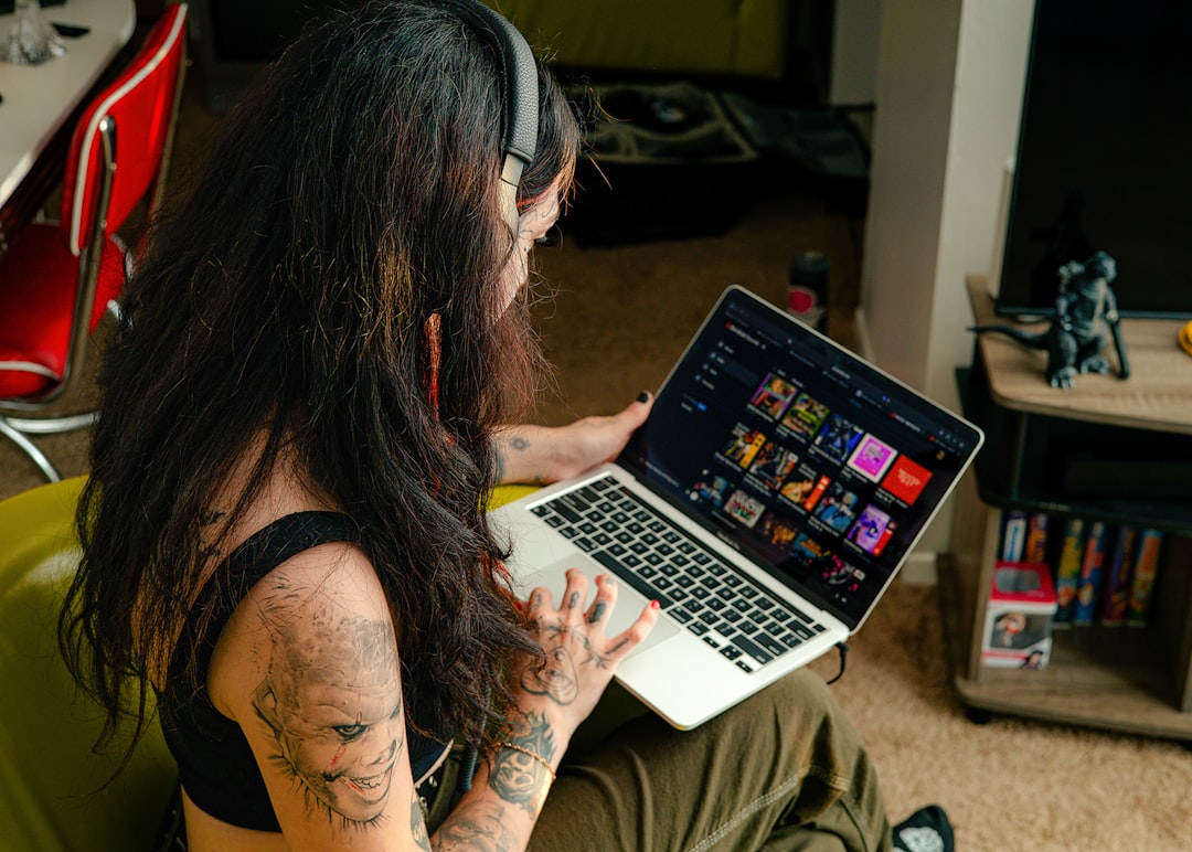 Person with long dark hair and visible tattoos, wearing headphones, using a laptop on their lap in a cozy living room.