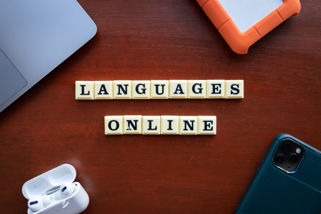 Scrabble tiles spell 'LANGUAGES ONLINE' on a wooden desk beside a laptop, earbuds case, tablet, and a smartphone nearby.