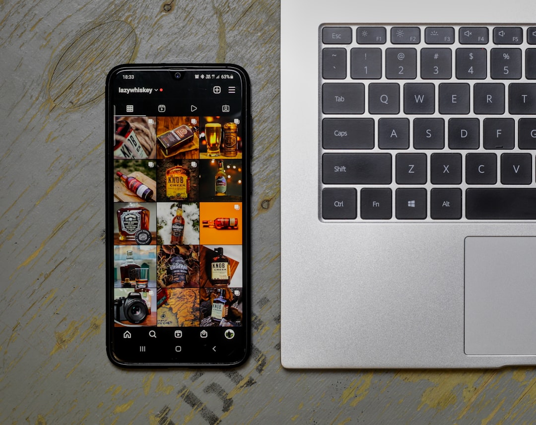 Smartphone on a wooden desk shows an Instagram grid of whiskey photos beside a silver laptop keyboard area to the right.