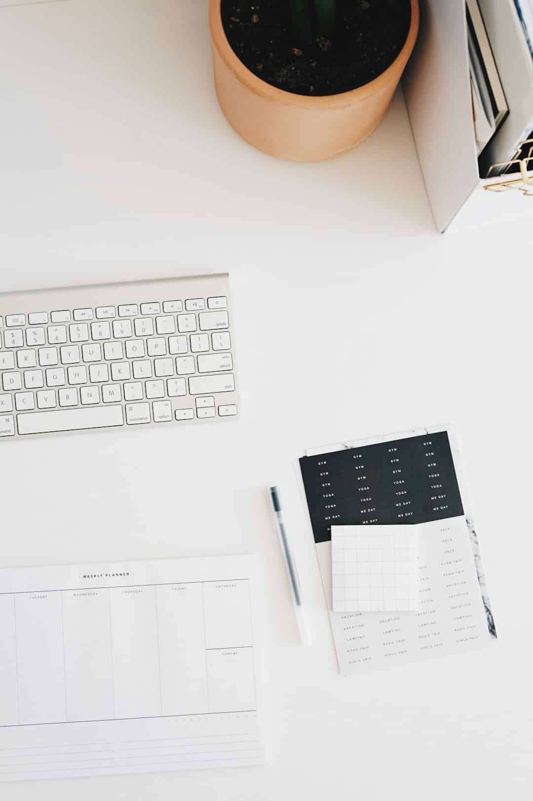 White desk workspace with a slim keyboard, planner sheets, a pen, and a potted plant viewed from above.