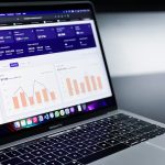 MacBook Pro displaying a dashboard with charts and metrics on a dark blue interface over a blurred background.