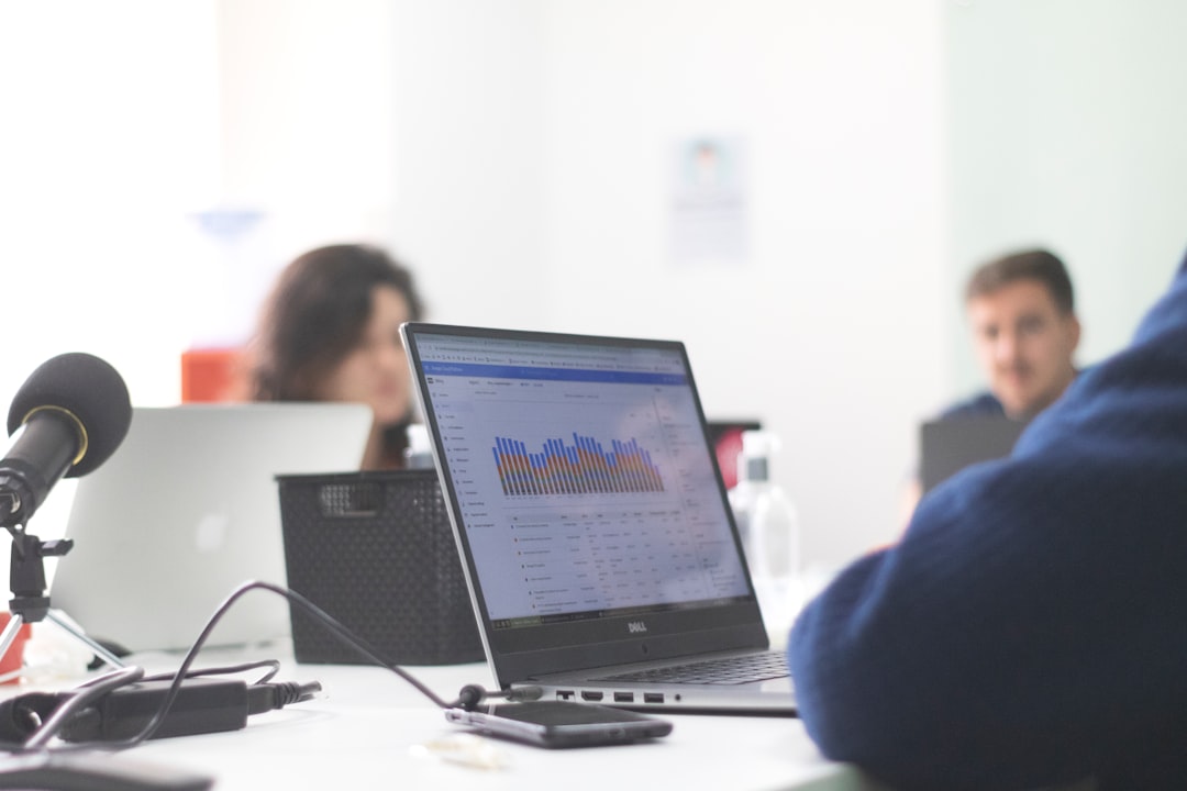 Conference desk with a laptop showing a colorful bar chart, a microphone in the foreground, and colleagues at work.