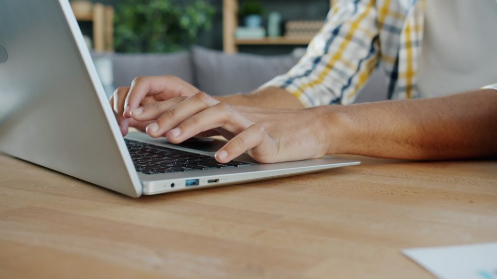Person typing on a silver laptop at a wooden desk in a cozy setting.