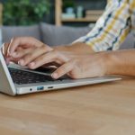 Person typing on a silver laptop at a wooden desk in a cozy setting.