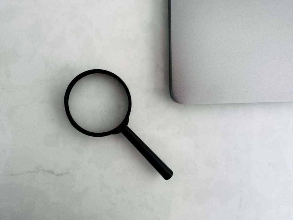 Magnifying glass sits near a laptop on a table keyword research tool search volume graph seo analytics workspace - AI Bud Black magnifying glass resting on a light marble desk beside a closed silver laptop, suggesting search or investigation