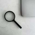 Black magnifying glass resting on a light marble desk beside a closed silver laptop, suggesting search or investigation
