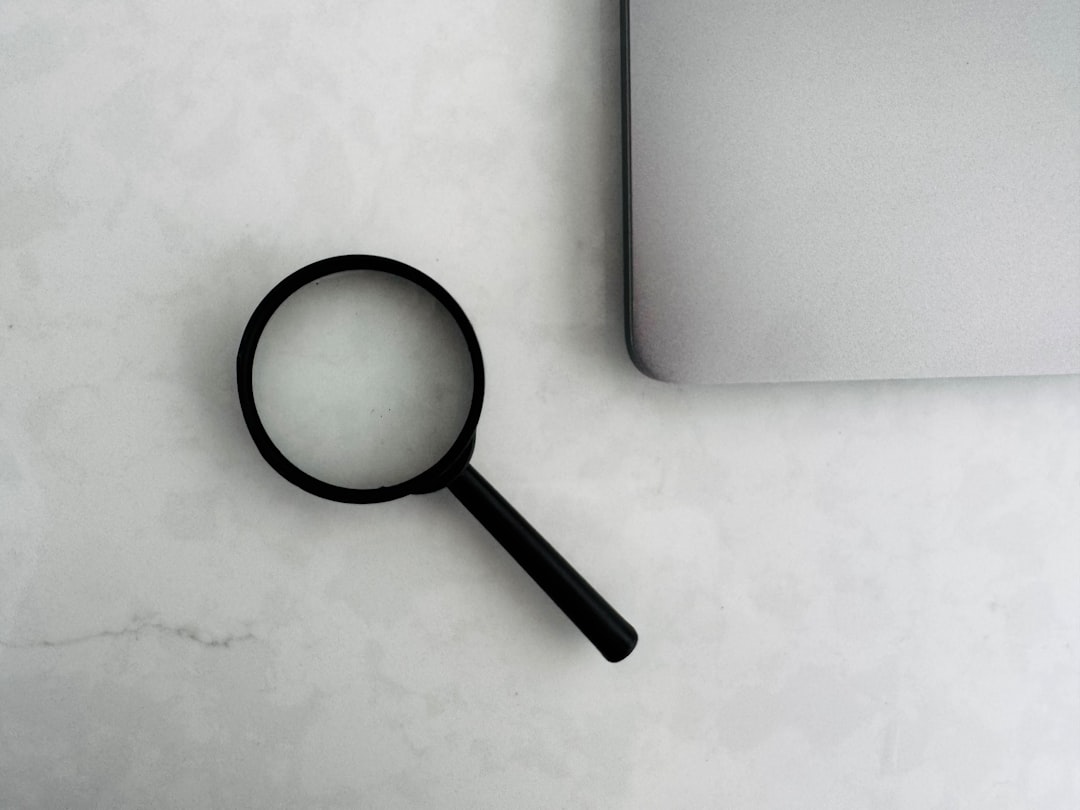 Black magnifying glass resting on a light marble desk beside a closed silver laptop, suggesting search or investigation