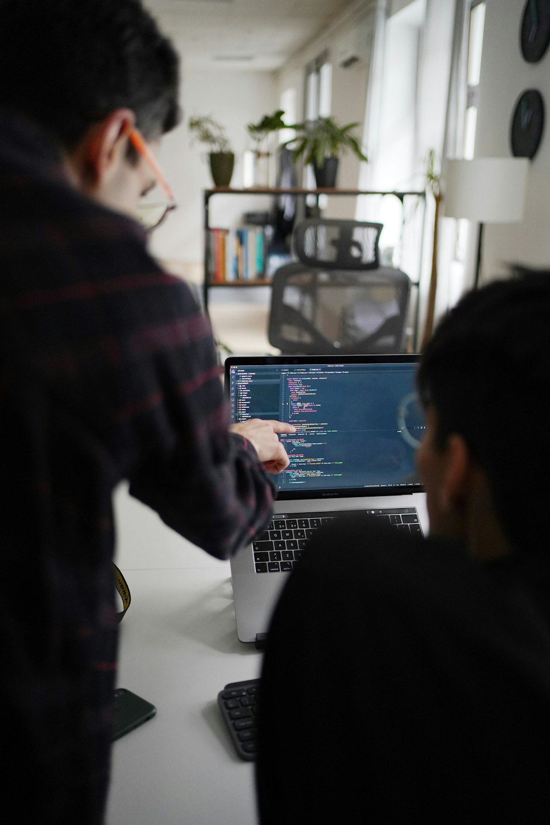 Two programmers collaborate at a laptop, one pointing at code on the screen in a bright office setting with plants in the background.