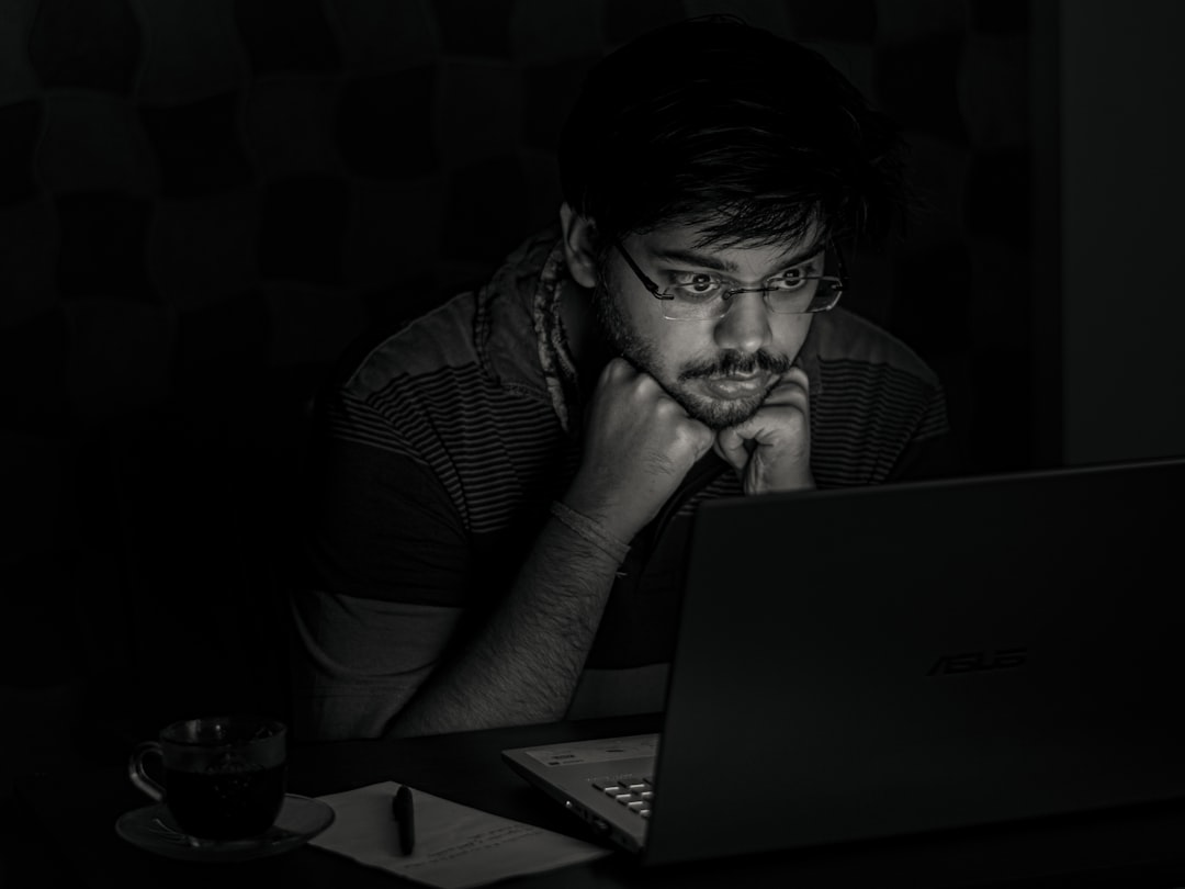 Young man with glasses sits at a desk in a dim room, chin resting on his hands as he looks at an open laptop.