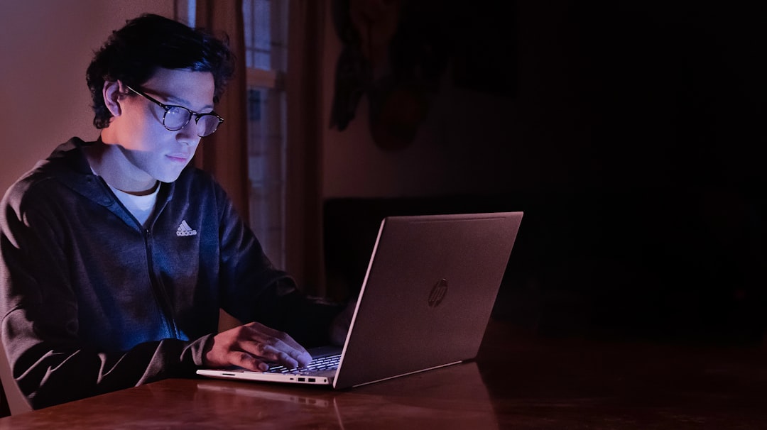Young person with glasses typing on a silver HP laptop at a wooden table in a dimly lit room with a blue-pink glow.