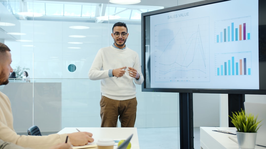 Man presenting sales data to a meeting, pointing to a large screen with line and bar charts labeled 'SALES VALUE' in a bright office conference room