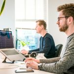 Two coworkers sit at bright desks in an open office, each typing on laptops with a monitor nearby and plants in the background.