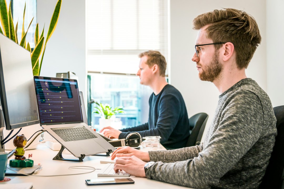 Two coworkers sit at bright desks in an open office, each typing on laptops with a monitor nearby and plants in the background.