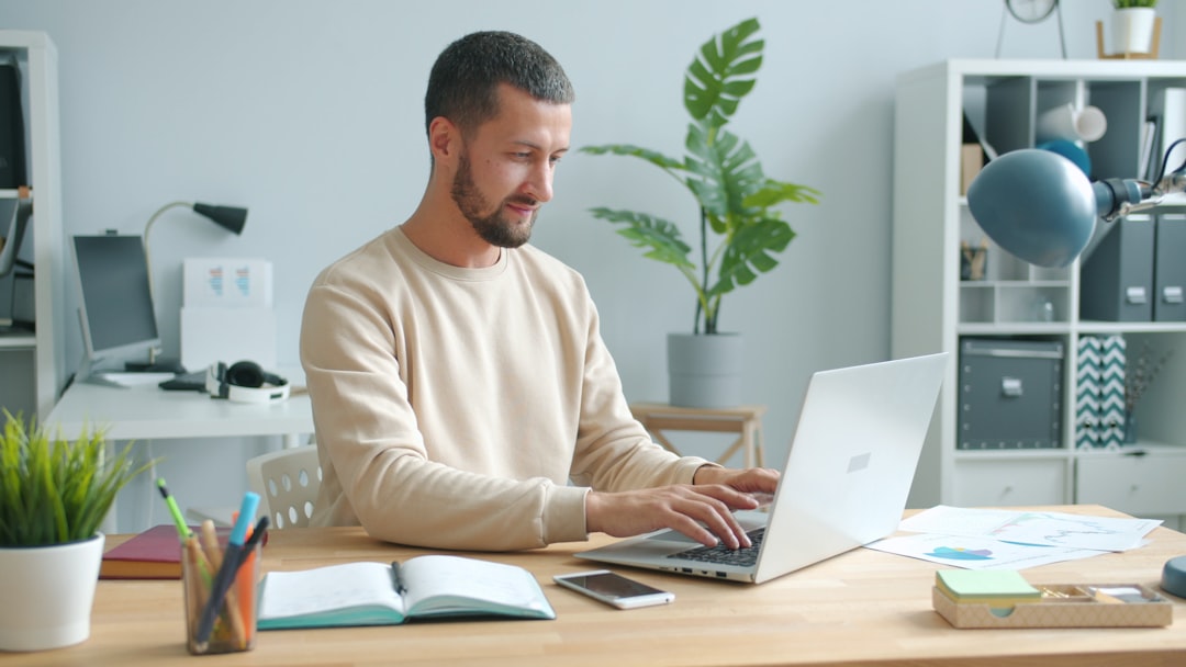 Man sits at a wooden desk typing on a silver laptop in a bright home office with plants and organized shelves in the background.