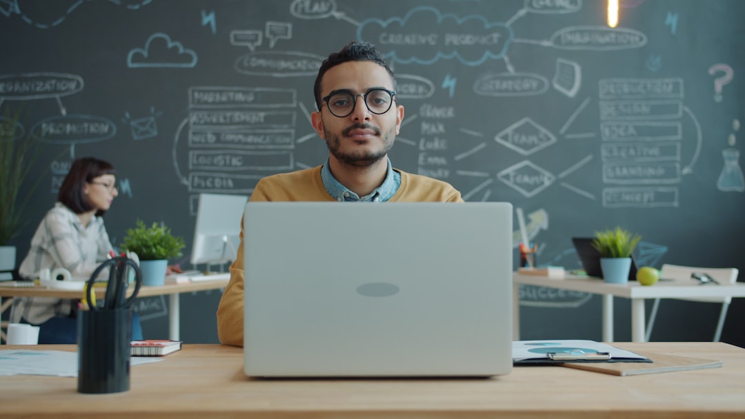 Man with glasses sits at a wooden desk working on a silver laptop, with a chalkboard full of diagrams behind him in a bright office setting.