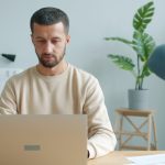 Man with short hair and beard in a beige sweatshirt sits at a wooden desk typing on a laptop in a bright, modern office with a large plant in the background