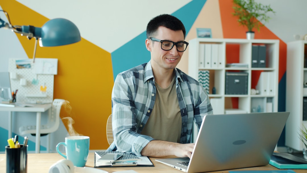 Man with glasses and a plaid shirt typing on a laptop at a bright modern office desk, steam rising from a blue mug nearby.