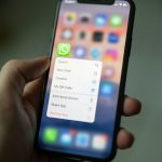 Hand holding an iPhone with WhatsApp options menu open: Search, New Chat, Camera, My QR Code, Share App, Remove App (blurred home screen)