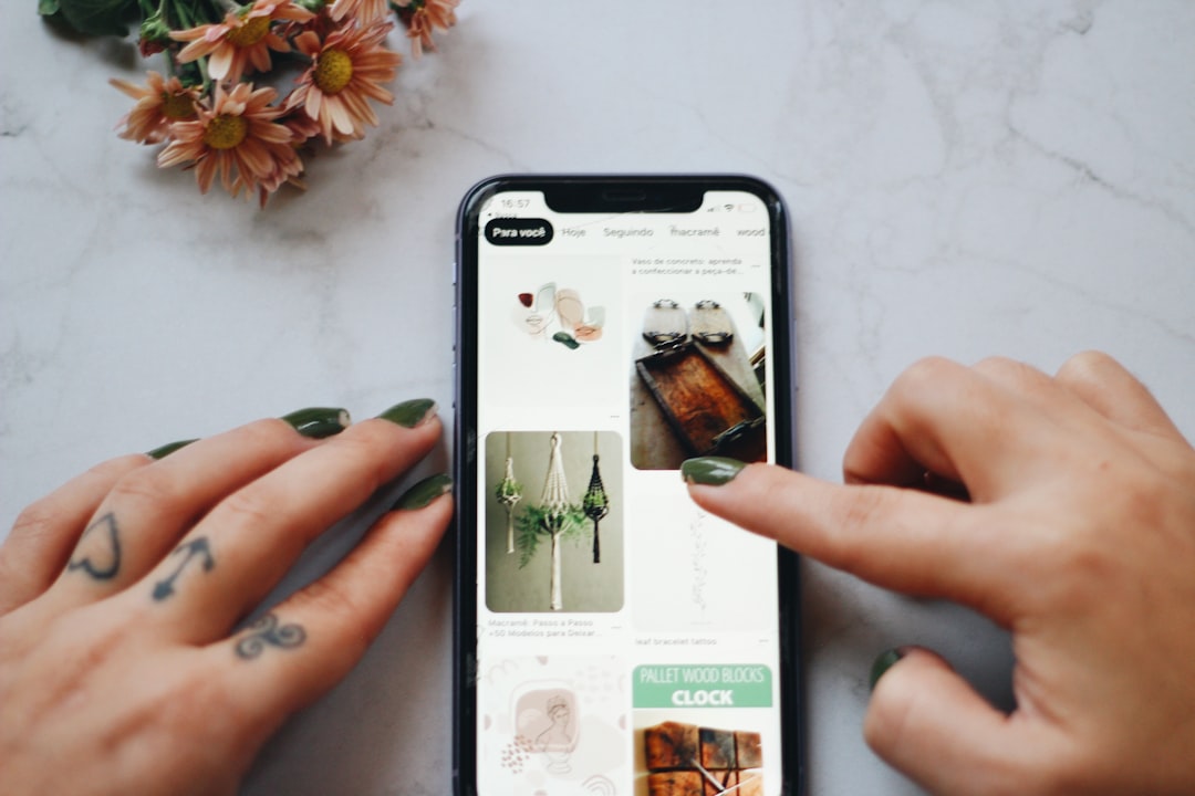 Close-up of hands with green nail polish scrolling through a phone screen displaying a grid of Pinterest-like pins, with a bouquet in the top-left corner.