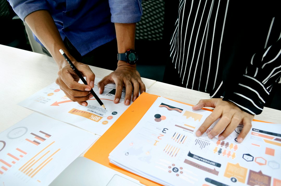 Two people review printed charts and graphs on a white table, one pointing with a pencil as they collaborate.