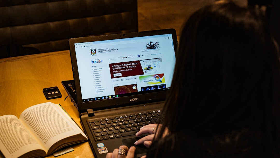 Person typing on an Acer laptop at a wooden table, with an open book beside it and a small device nearby.