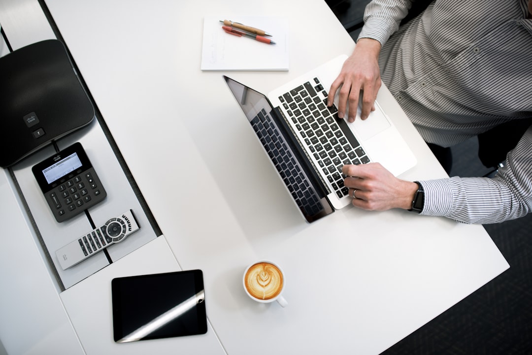 Person typing on a laptop at a white desk, with a coffee cup, tablet, and a safe nearby on the surface.