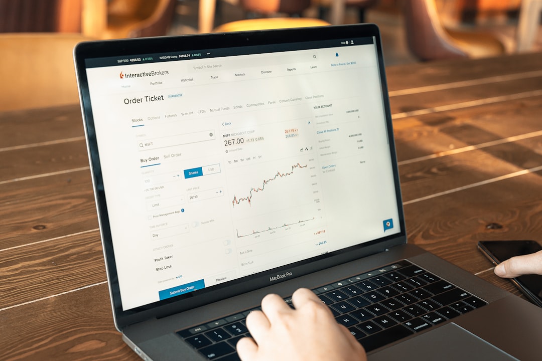 Person using a MacBook Pro to view an Interactive Brokers order ticket and stock chart on a wooden table.