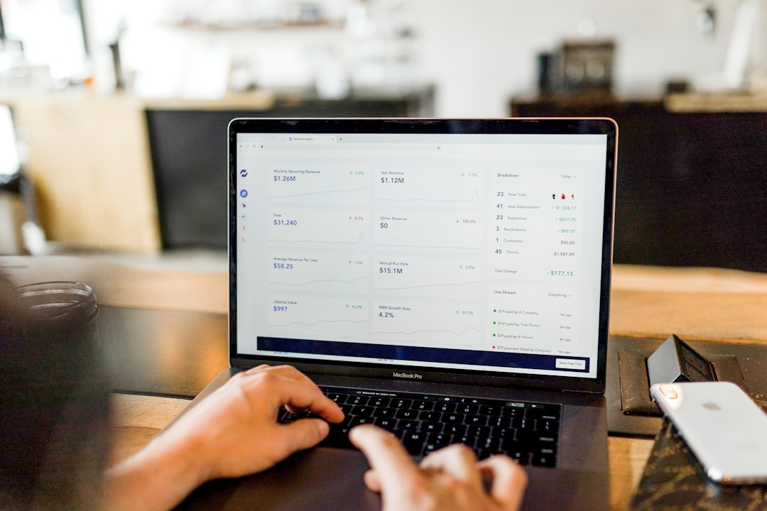 Person typing on a MacBook Pro displaying a business analytics dashboard on a wooden table in a bright, casual workspace.