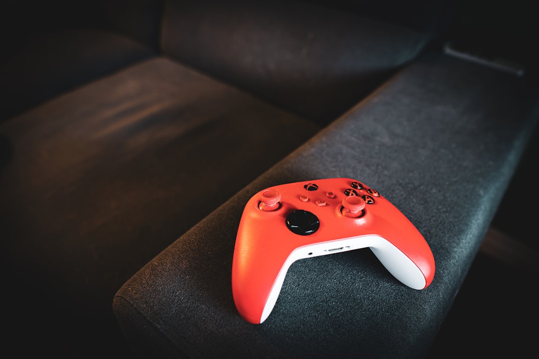 Red wireless game controller resting on a dark sofa arm in dim lighting.