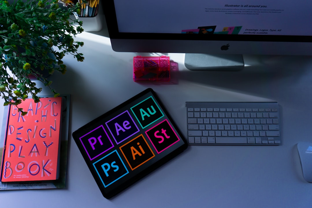 Top-down desk scene with a tablet displaying Adobe app icons, a keyboard, monitor, plant, and pink storage box.