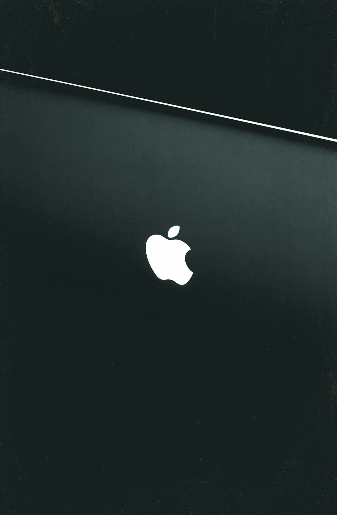 White Apple logo on a dark textured surface with a diagonal light line across the top-left corner, minimal and brand-focused.