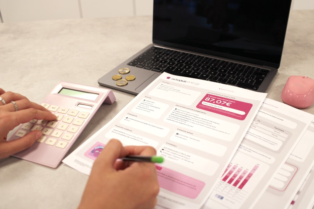 Person using a pink calculator and writing on financial documents at a gray desk, with coins, a laptop, and a pink mouse nearby.