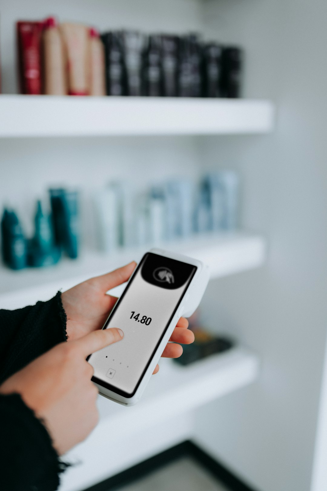 Person holding a white handheld payment terminal showing a total of 14.80 in a store with blurred shelves in the background