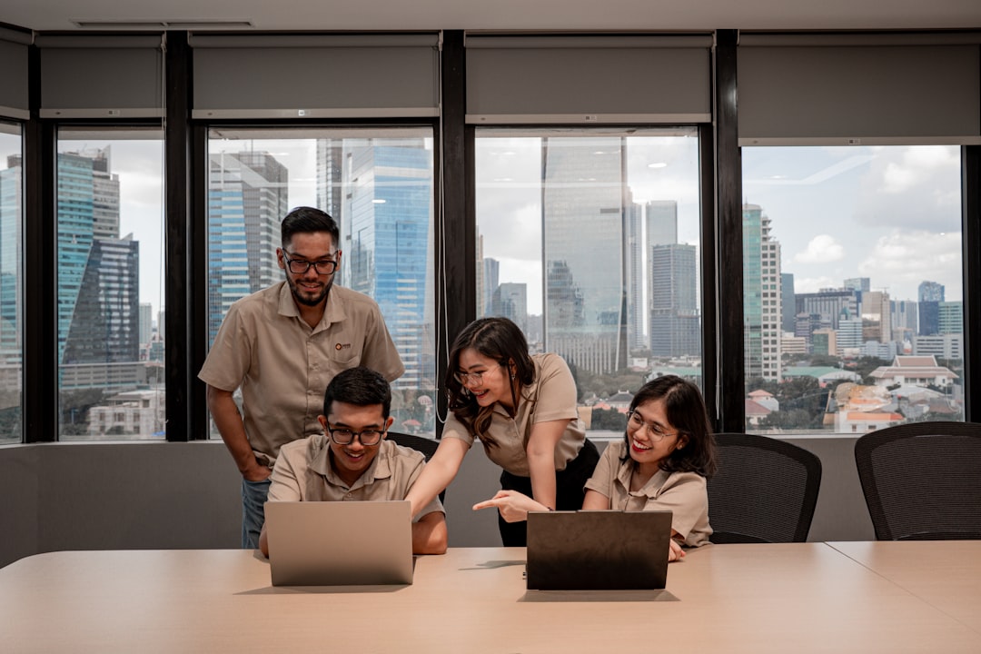 Four coworkers in beige shirts collaborating around laptops at a conference table with a city skyline visible outside the windows.