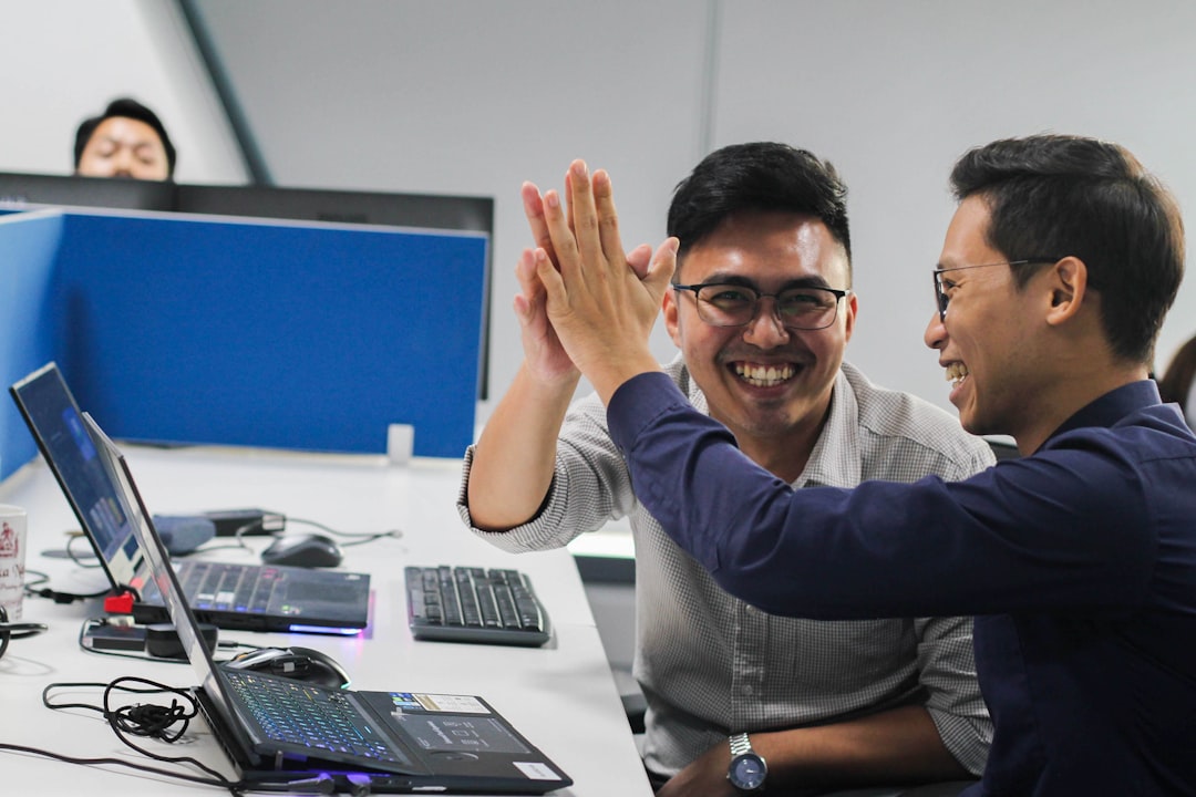 Two coworkers share a high-five and smile at a desk with laptops and monitors in an office.