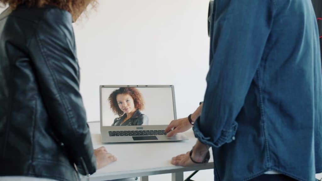 Two people stand at a table, looking at a laptop screen showing a woman during a video call or virtual meeting