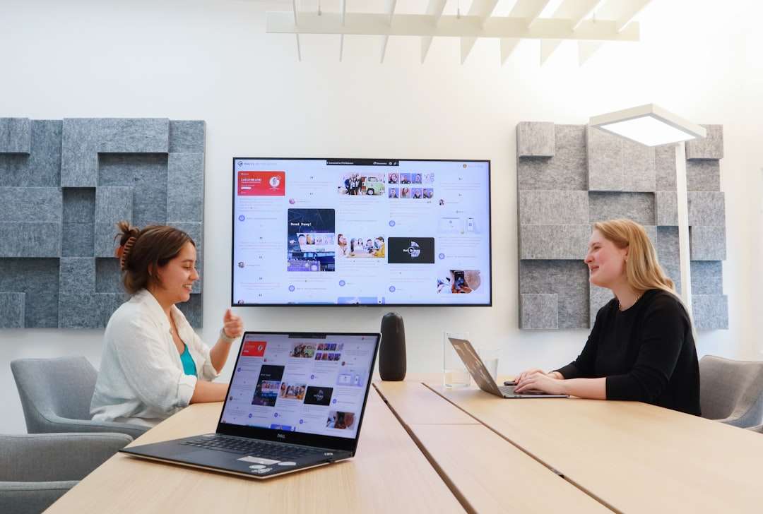 Two women sit at a wooden table with laptops; a large wall screen shows a social media page.