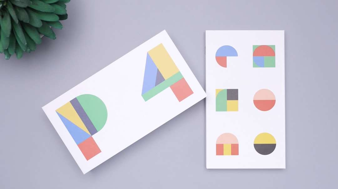 Two white cards with colorful geometric shapes forming abstract letters, placed on a gray background near a green plant corner.