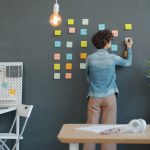 Person in a denim shirt sticks notes on a chalkboard wall for a planning session in a bright, organized office.