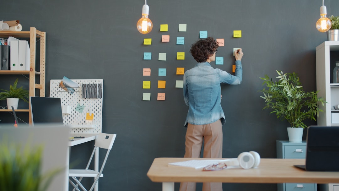 Person in a denim shirt sticks notes on a chalkboard wall for a planning session in a bright, organized office.