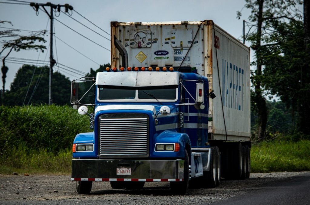 Front view of a blue semi-truck with a chrome grille hauling a large white refrigerated container along a rural road at the edge of green fields.