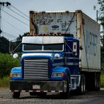 Front view of a blue semi-truck with a chrome grille hauling a large white refrigerated container along a rural road at the edge of green fields.