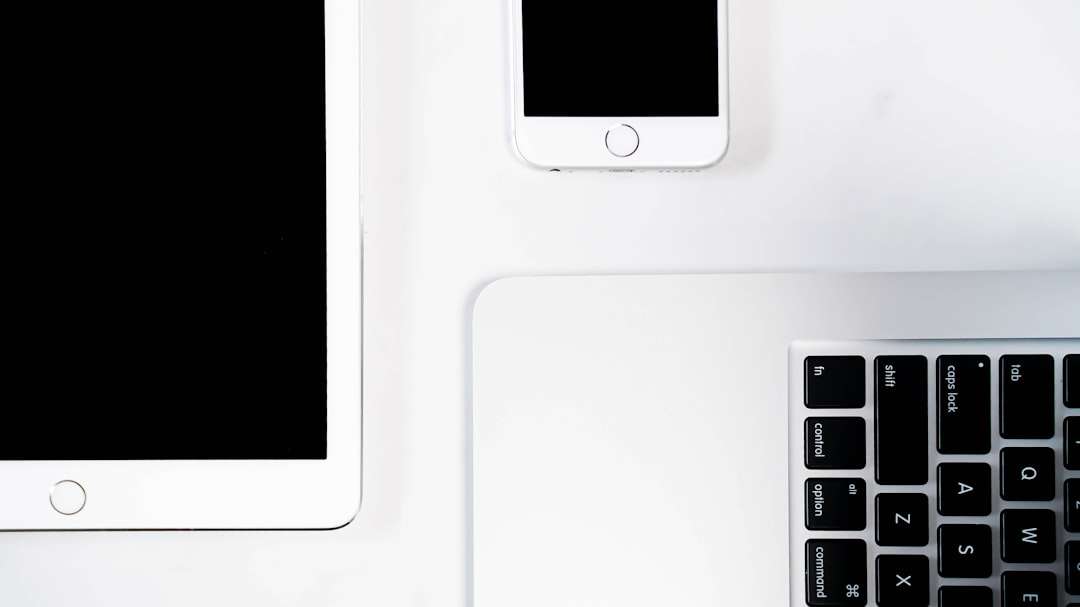 Overhead view of a white tablet on the left, a white iPhone above it, and a silver laptop keyboard on a white desk.