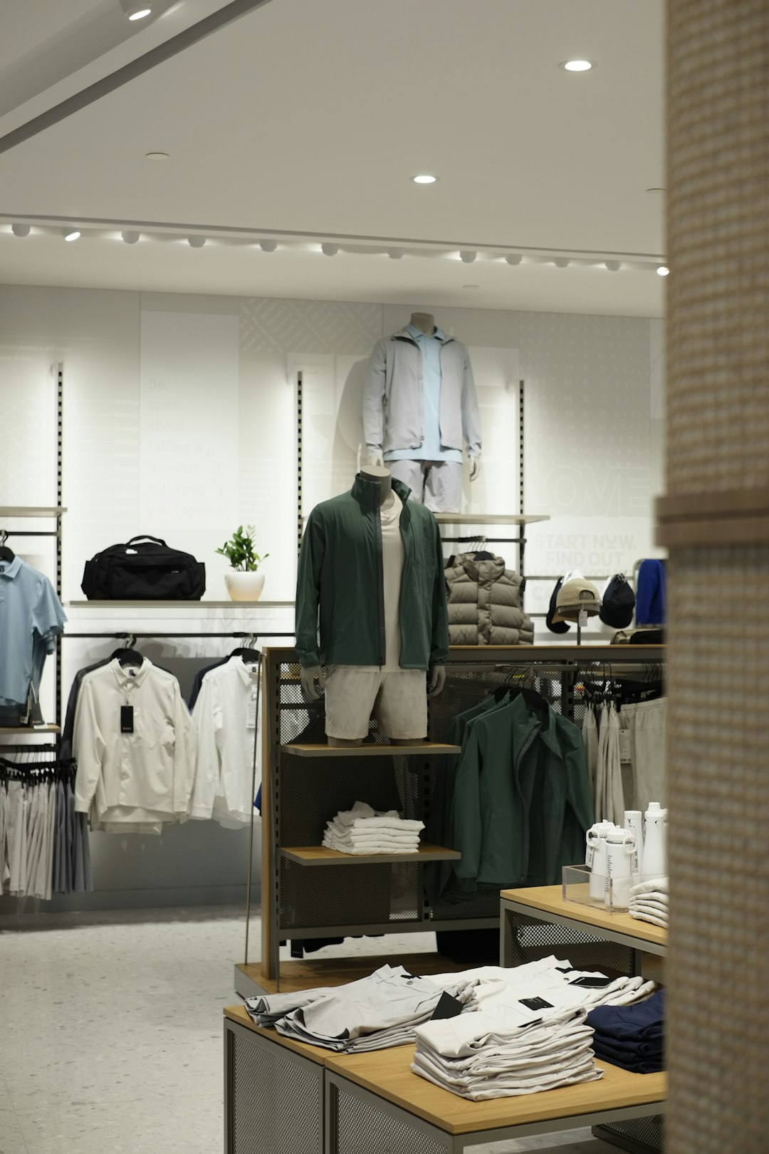 Bright clothing store display with mannequins wearing jackets and casual wear, surrounded by folded shirts and racks of apparel in a well-lit shop.